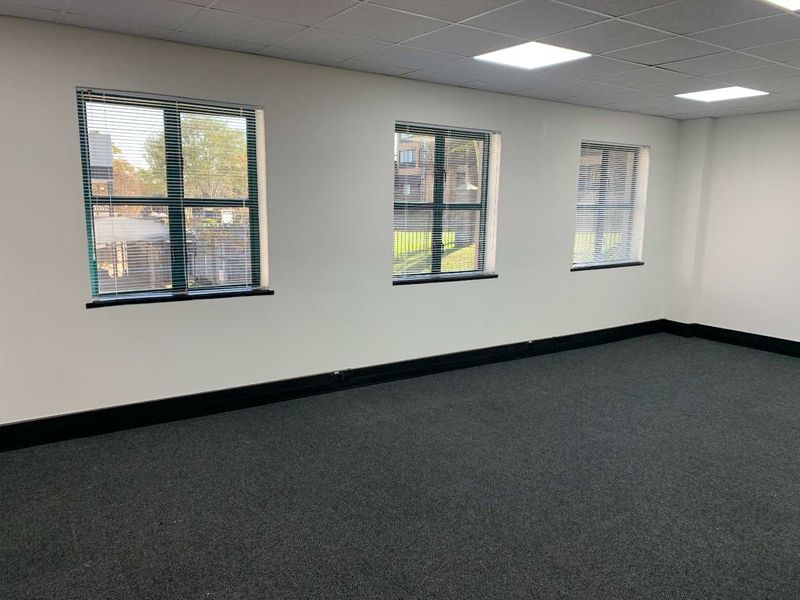 A bright, vacant office space featuring neutral walls, dark carpeting, suspended ceiling with recessed lighting, and multiple windows with fitted venetian blinds.