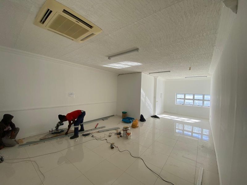 A spacious, empty office area featuring tiled flooring, recessed ceiling air conditioning, and natural light from a large window.