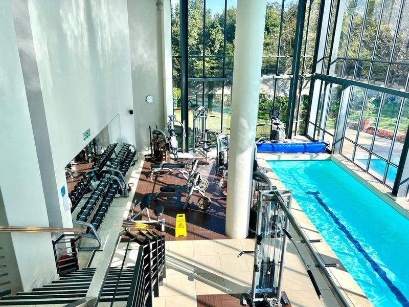 A modern indoor gym facility featuring a lap pool, floor-to-ceiling windows with garden views, and a mirrored wall, providing a bright and spacious workout environment.