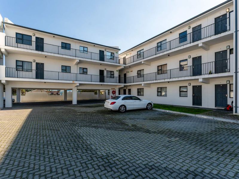 Exterior view of a multi-story apartment complex featuring paved parking area, external walkways with metal railings, and multiple unit entrances.