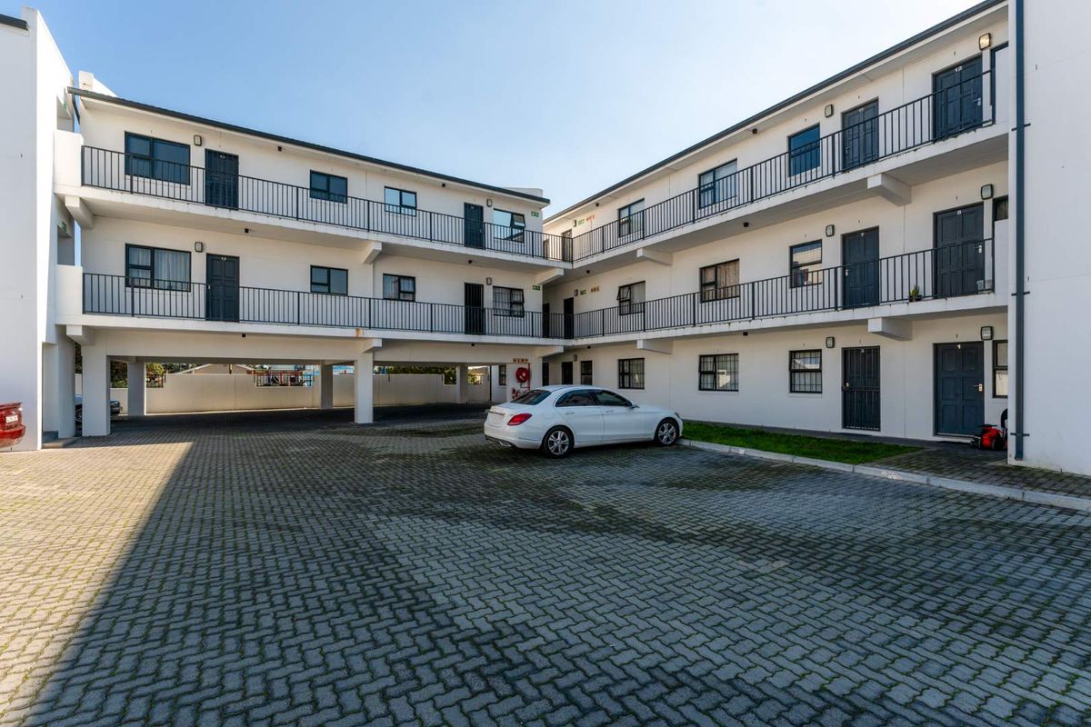 Exterior view of a multi-story apartment complex featuring paved parking area, external walkways with metal railings, and multiple unit entrances.