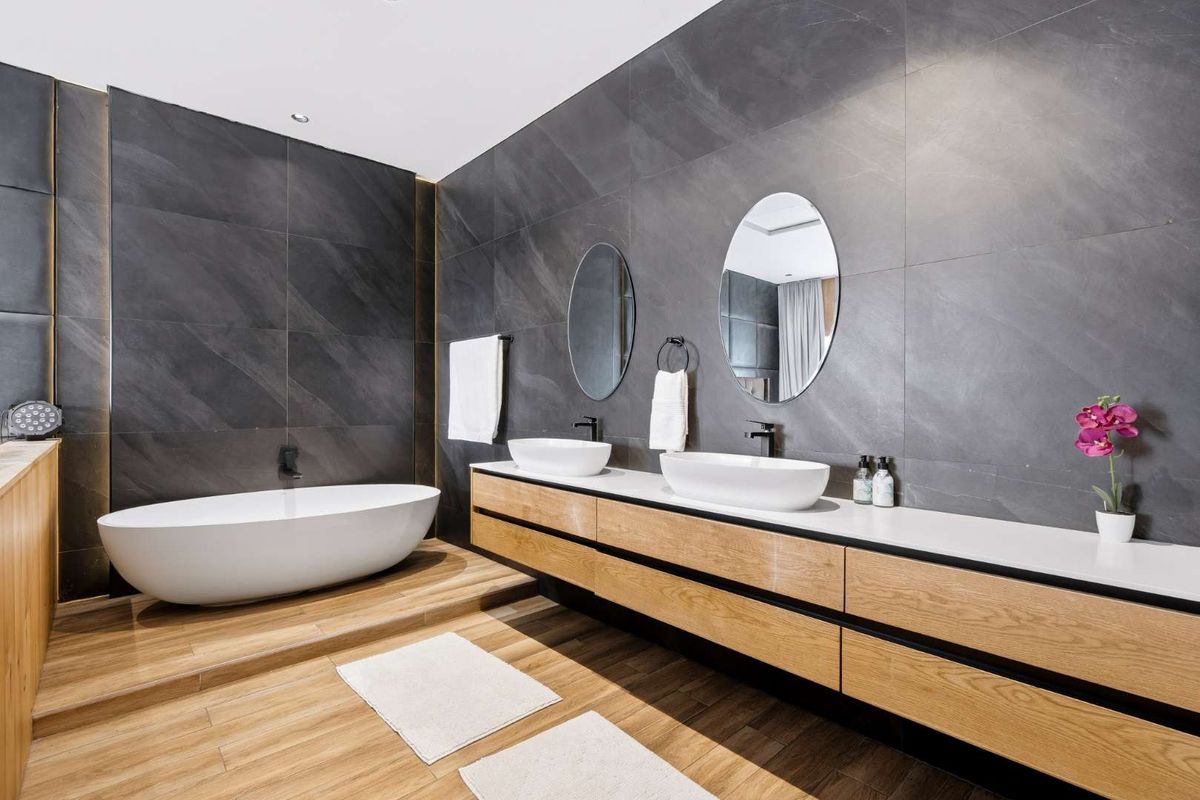 Modern luxury bathroom featuring a freestanding bathtub, double vessel sink vanity with timber cabinetry, and dark large-format wall tiling.