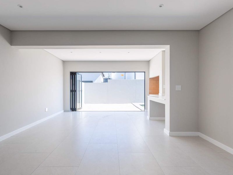 Spacious open-plan living area featuring neutral tiled flooring, recessed lighting, and a built-in braai, with seamless access to the outdoor patio through stacking doors.