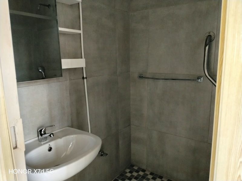 A modern bathroom featuring grey wall tiles, a pedestal basin with chrome mixer tap, and a shower area equipped with a safety grab rail.
