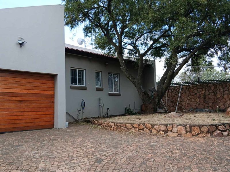Front exterior view featuring a paved driveway, a wooden garage door, and a stone retaining wall with established trees.