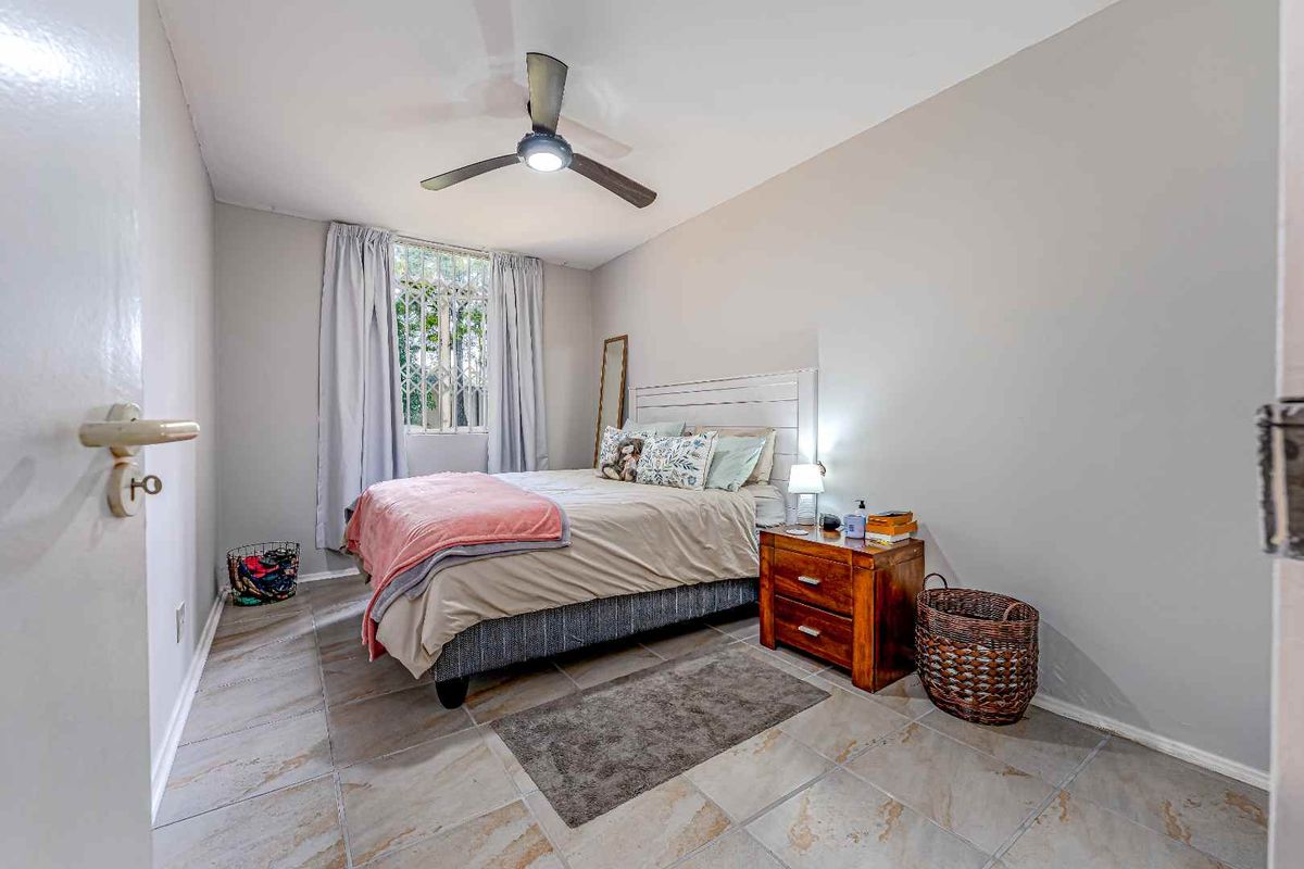 A functional bedroom featuring tiled flooring, a ceiling fan, and a window fitted with security bars.