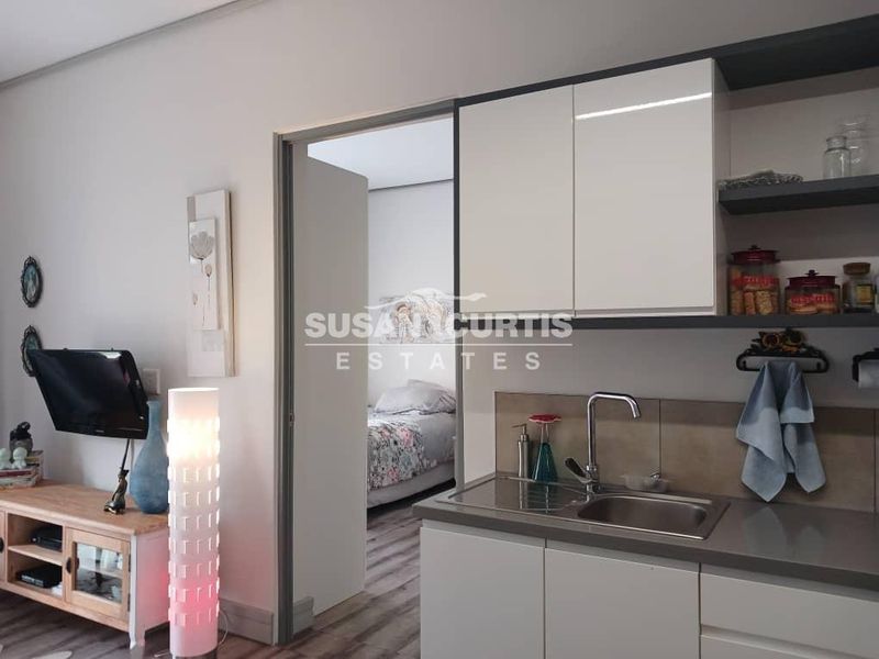 A compact kitchen area featuring modern white cabinetry, stone benchtops, and a stainless steel sink, with an adjacent view into a bedroom.