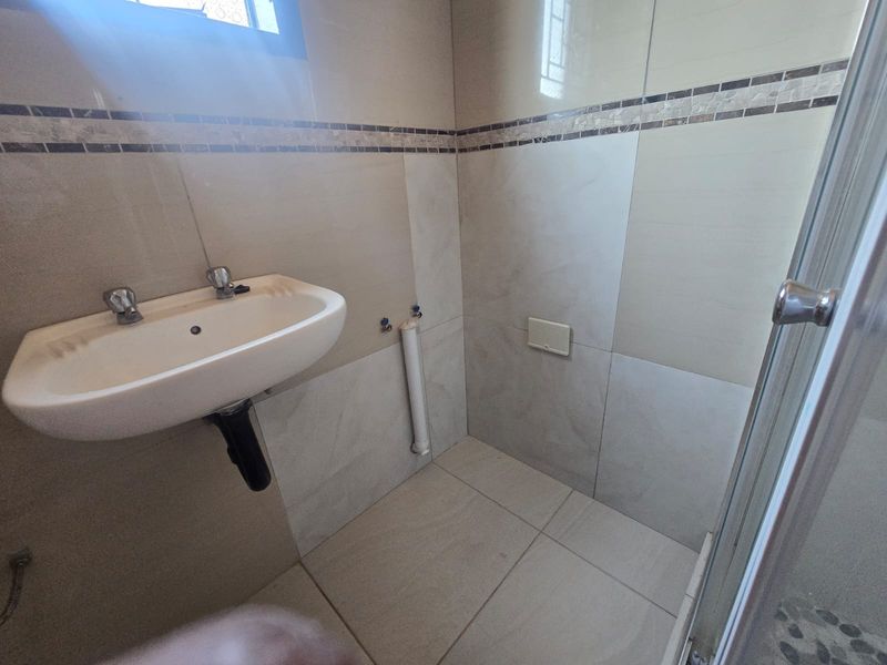 A functional bathroom featuring a wall-mounted basin, shower enclosure, and neutral ceramic tiling throughout. The space is ready for vanity installation.