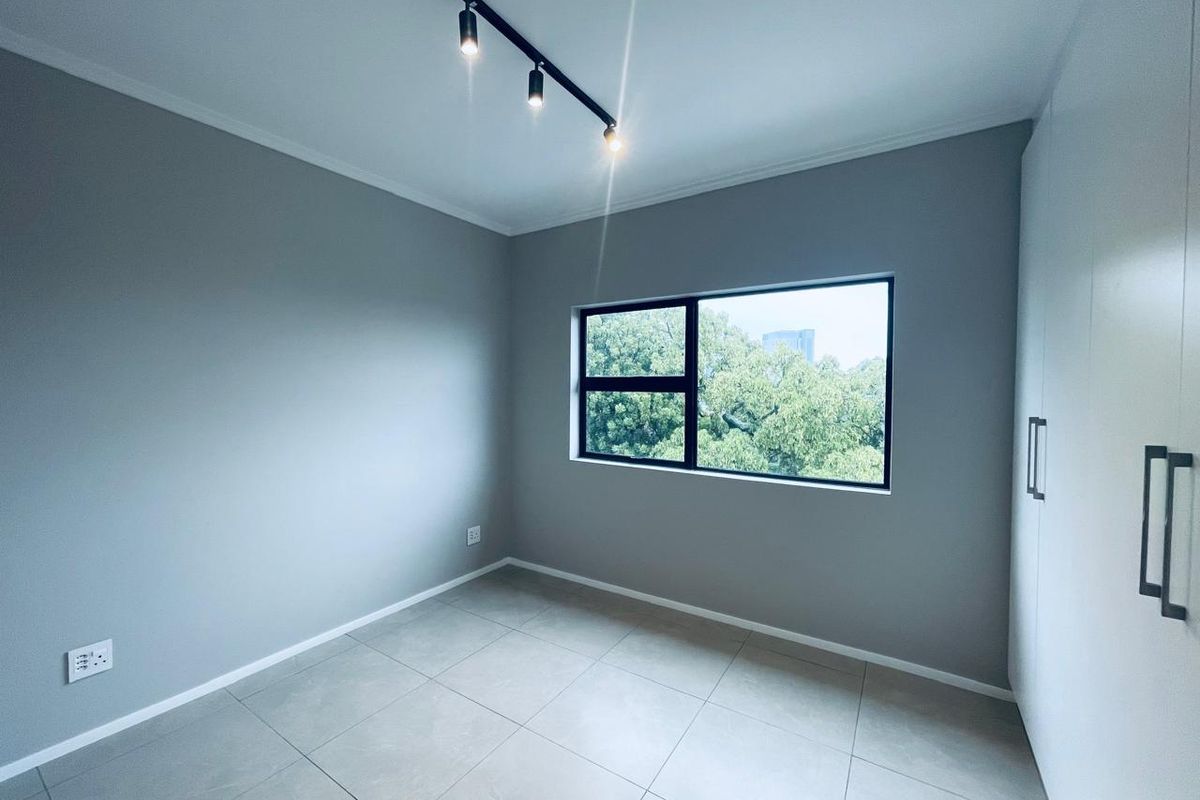 A bright, modern bedroom featuring neutral grey walls, large format floor tiles, built-in storage, and contemporary track lighting with a pleasant garden view.