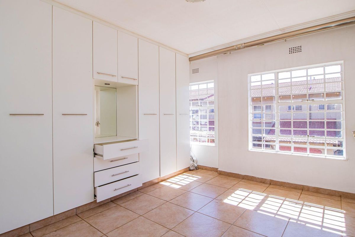 A bright bedroom featuring extensive built-in white cabinetry with integrated vanity mirror, tiled flooring, and large windows allowing for ample natural light.