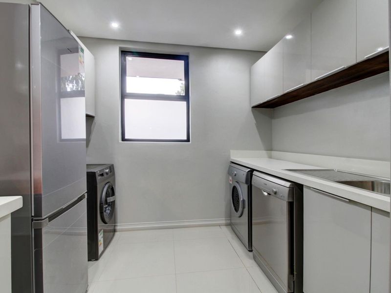 A modern, well-appointed laundry room featuring sleek white cabinetry, stone countertops, a stainless steel sink, and integrated appliance spaces with recessed lighting.