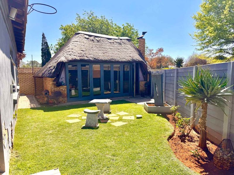 A well-maintained rear garden featuring a thatched-roof structure with folding glass doors, a brick chimney, and a modern water feature.