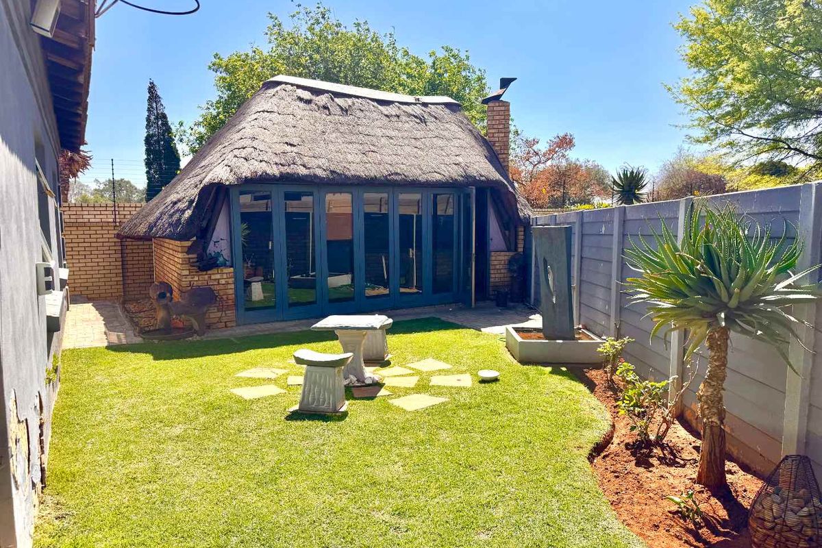 A well-maintained rear garden featuring a thatched-roof structure with folding glass doors, a brick chimney, and a modern water feature.