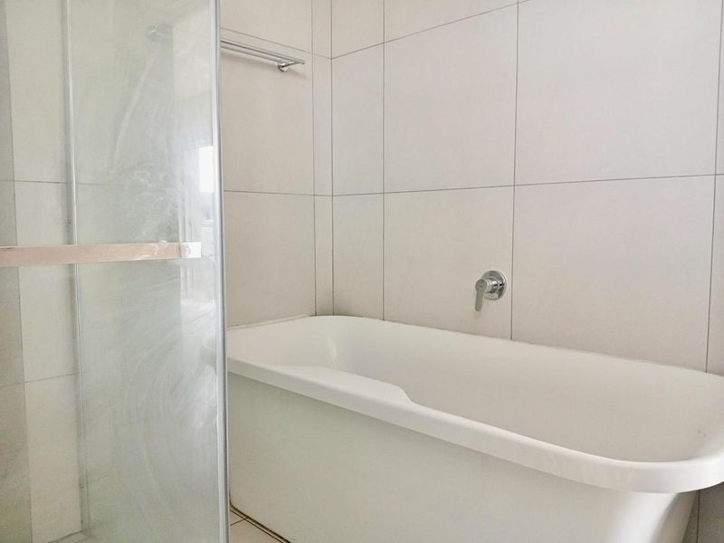 A clean, modern bathroom featuring a white bathtub, glass shower screen, and neutral wall tiling with chrome fixtures.
