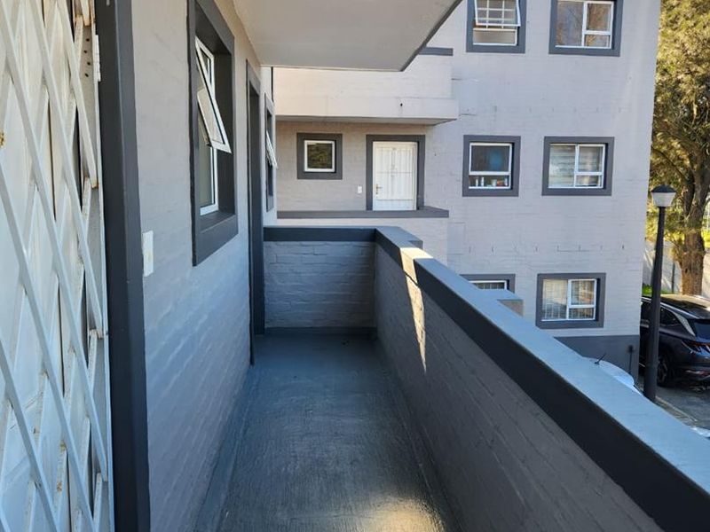 A clean, covered balcony area featuring painted brick walls, concrete flooring, and a security gate, overlooking the apartment complex grounds.