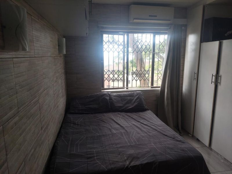 A compact bedroom featuring built-in wardrobes, wall-mounted air conditioning, and tiled wall finishes, with a security gate over the window.