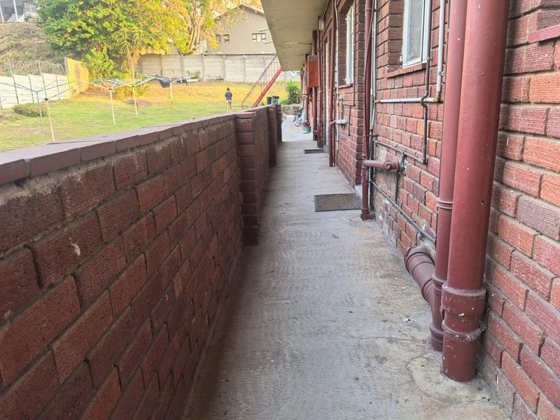 A narrow exterior rear walkway featuring exposed brick walls, concrete flooring, and visible plumbing infrastructure leading towards a communal garden area.