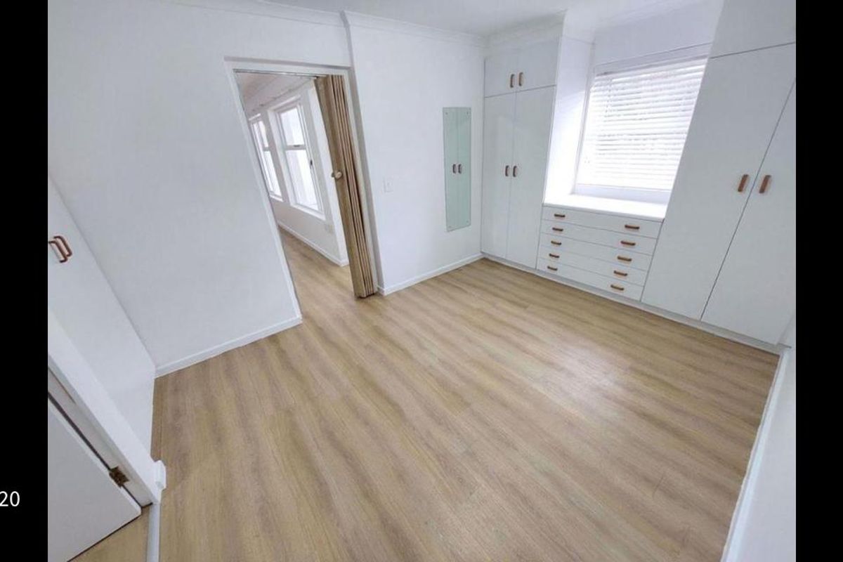 A bright, empty bedroom featuring light wood-look flooring, extensive built-in cabinetry with integrated drawers, and a window with fitted blinds.