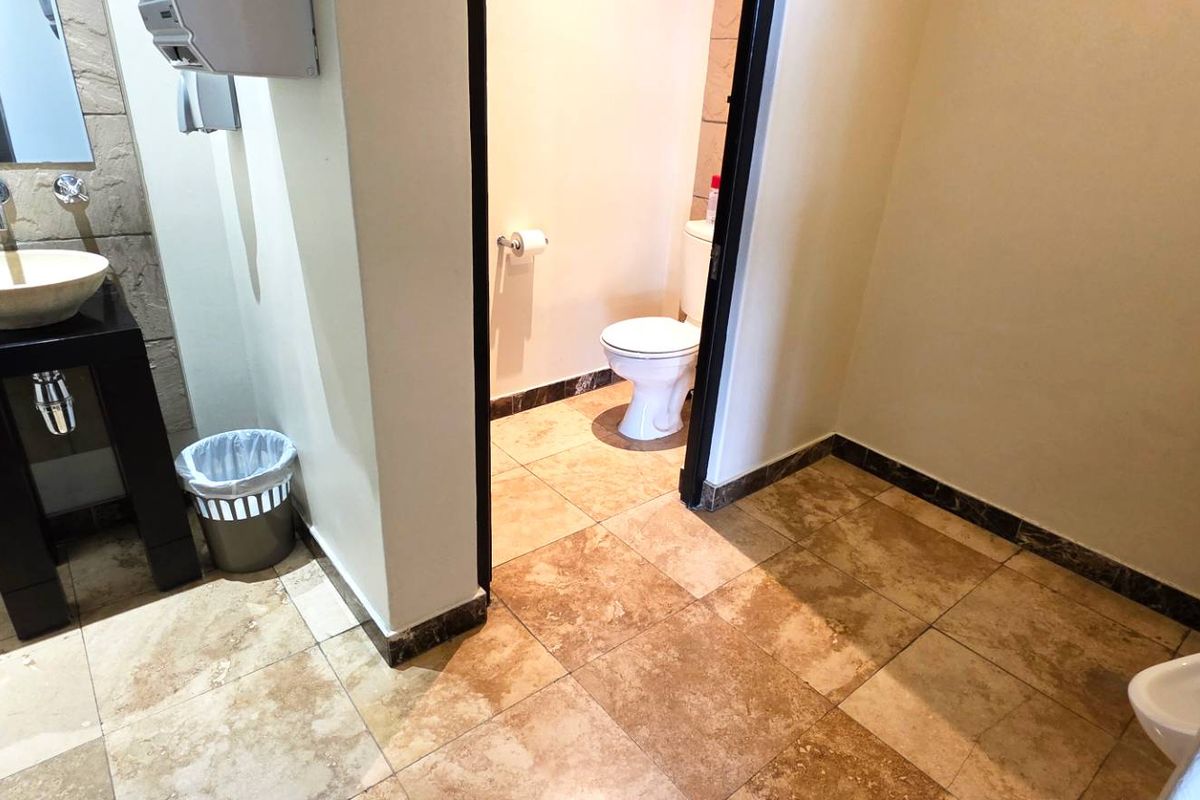 A modern office restroom featuring a stone vessel sink, tiled flooring, and a separate enclosed toilet cubicle.