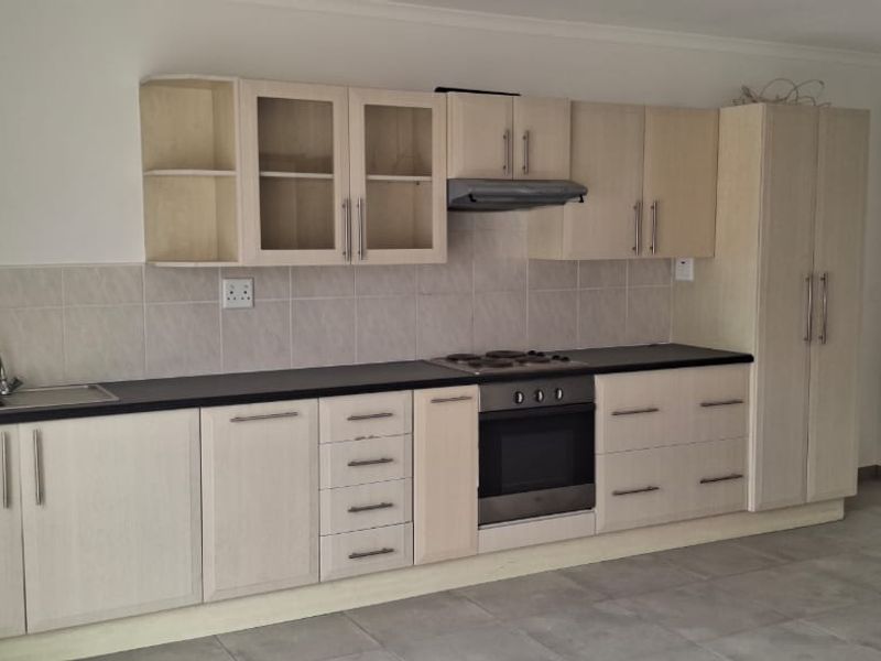 A clean, functional kitchen featuring light wood-finish cabinetry, dark countertops, integrated appliances, and neutral tiled flooring, offering a bright and practical workspace.