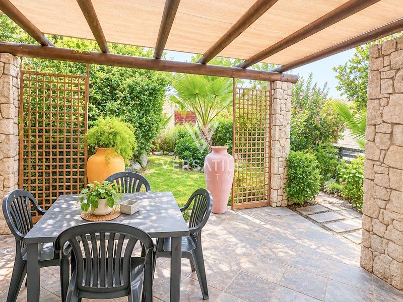 A covered outdoor patio featuring stone-clad pillars, a timber pergola with a shade sail, and tiled flooring, overlooking a landscaped garden.
