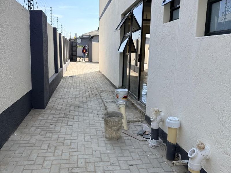A side exterior walkway featuring paved flooring, boundary wall with electric fencing, and exposed plumbing infrastructure along the building facade.