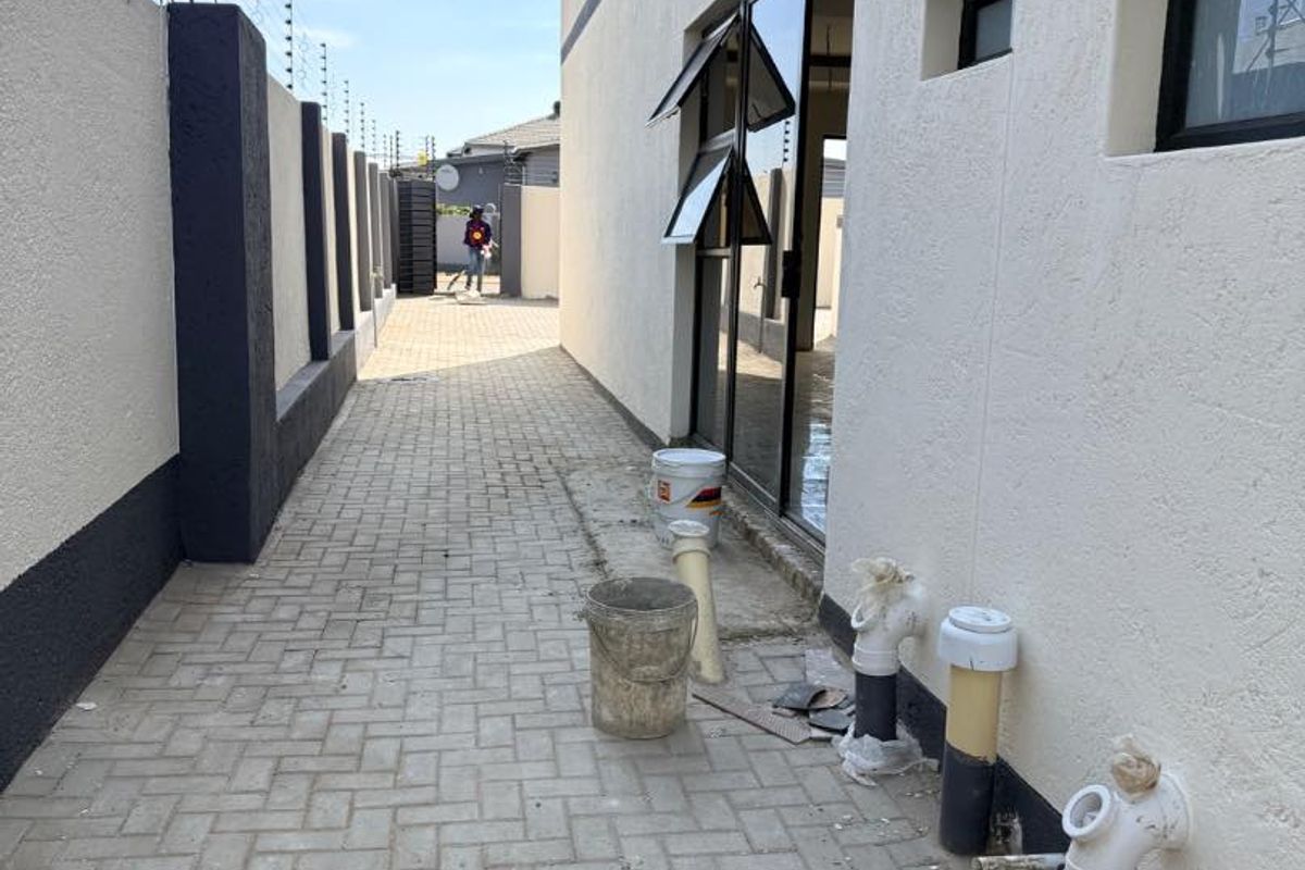 A side exterior walkway featuring paved flooring, boundary wall with electric fencing, and exposed plumbing infrastructure along the building facade.