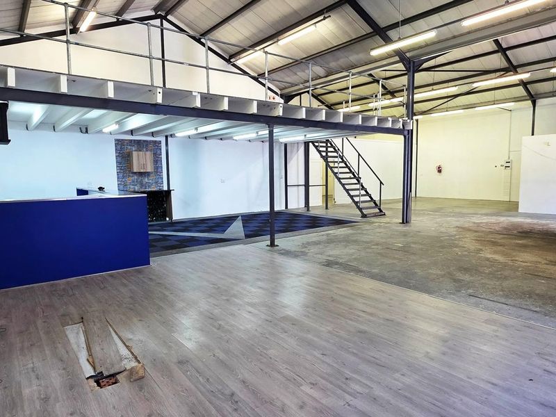 Spacious industrial utility area featuring a mezzanine level, steel staircase, exposed roof trusses, and a combination of laminate and concrete flooring.