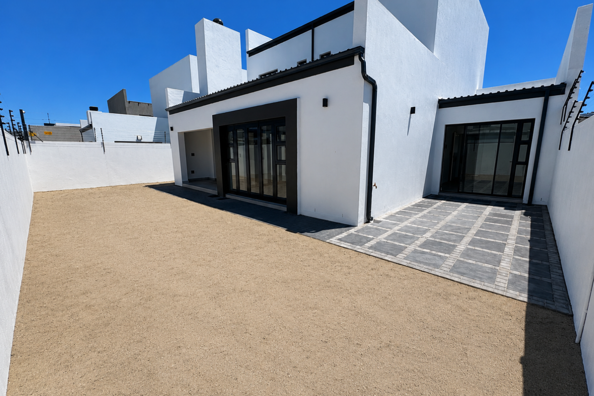 A modern rear exterior view featuring a paved patio area, white rendered walls, black window frames, and a large, low-maintenance gravel garden space.