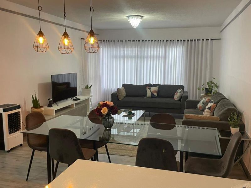A well-lit open-plan living and dining area featuring modern pendant lighting, neutral walls, and laminate flooring with large windows covered by vertical blinds.