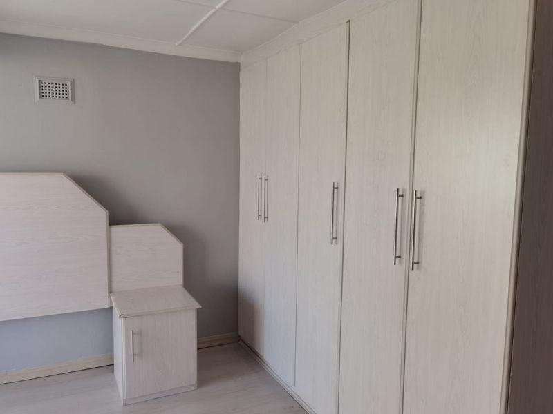 A clean bedroom featuring light wood-grain built-in cupboards and a matching integrated headboard unit with neutral grey walls.