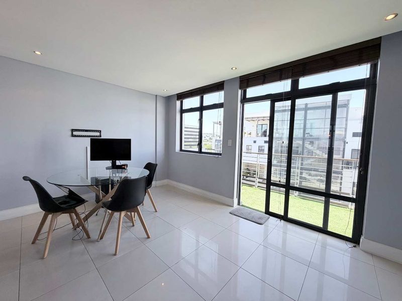 Bright living area featuring large aluminium framed windows and sliding doors leading to a balcony, finished with modern tiled flooring and recessed lighting.