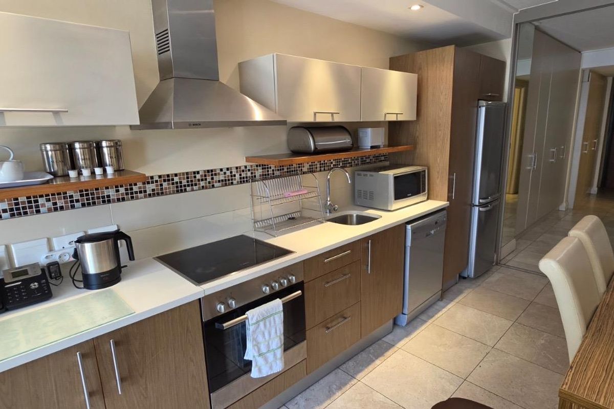 A modern galley-style kitchen featuring wood-finish cabinetry, stainless steel appliances, a mosaic tile backsplash, and tiled flooring, seamlessly integrated into the open-plan living space.