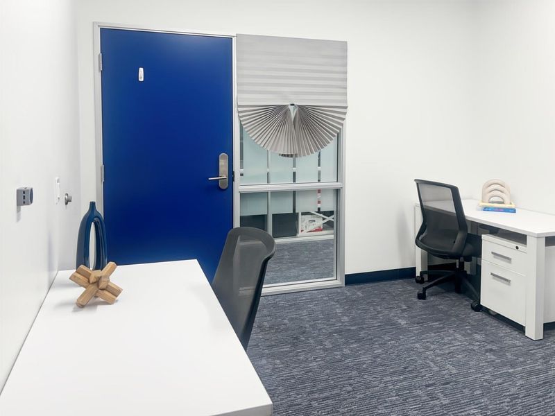 A clean, modern office space featuring carpeted flooring, a blue entrance door, and a glass partition with a pleated blind.