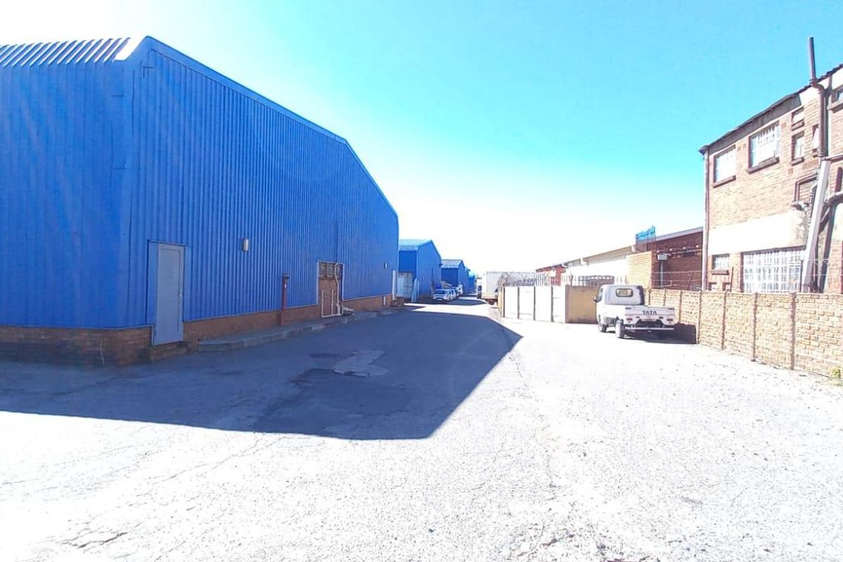 Industrial complex exterior featuring blue corrugated metal warehouse structures, paved access driveway, and brick perimeter walling.