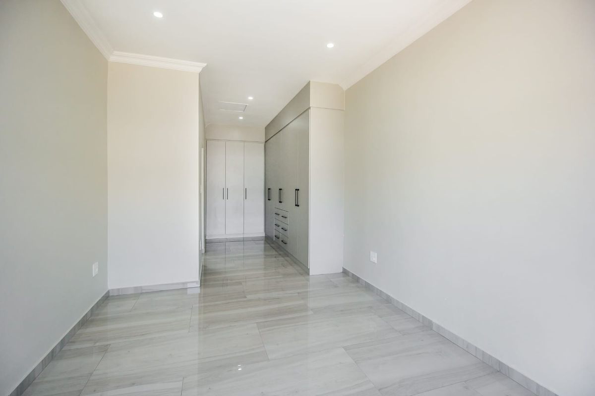 A bright, empty bedroom featuring modern grey tiled flooring, neutral walls, and extensive built-in cupboard storage with recessed downlighting.