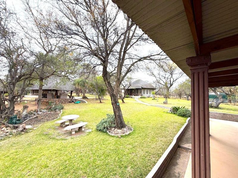 A covered patio area overlooking a spacious garden with mature trees and additional outbuildings in the background.
