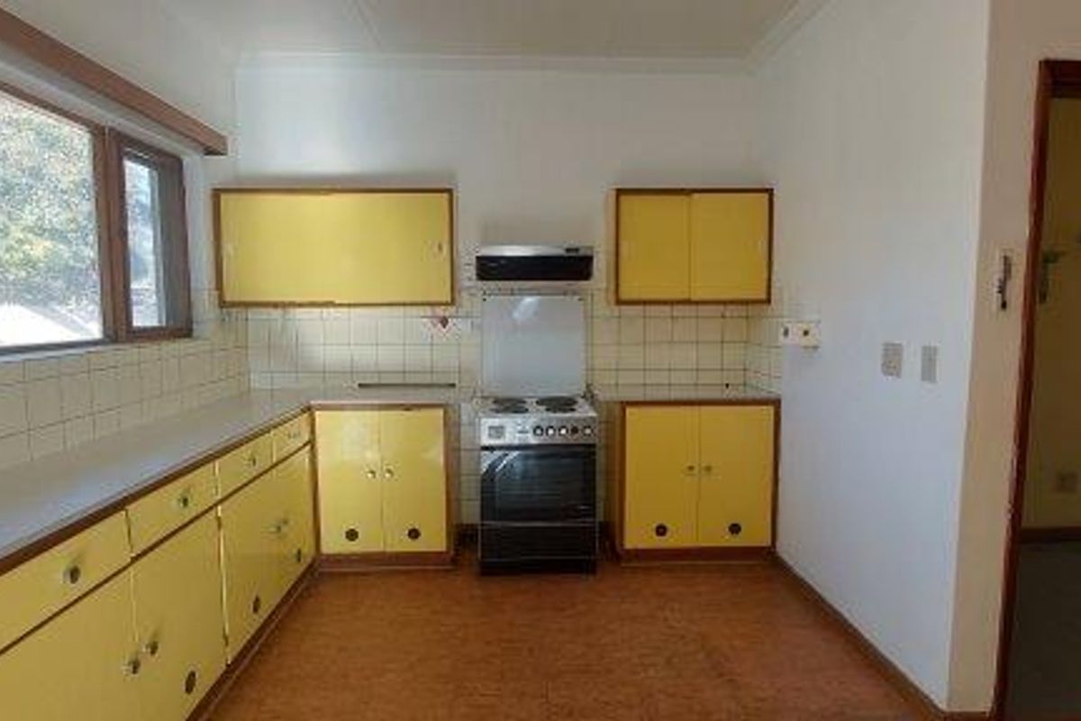 A dated kitchen featuring yellow cabinetry, a freestanding stove with extractor fan, and tiled splashbacks, offering a functional layout with natural light.