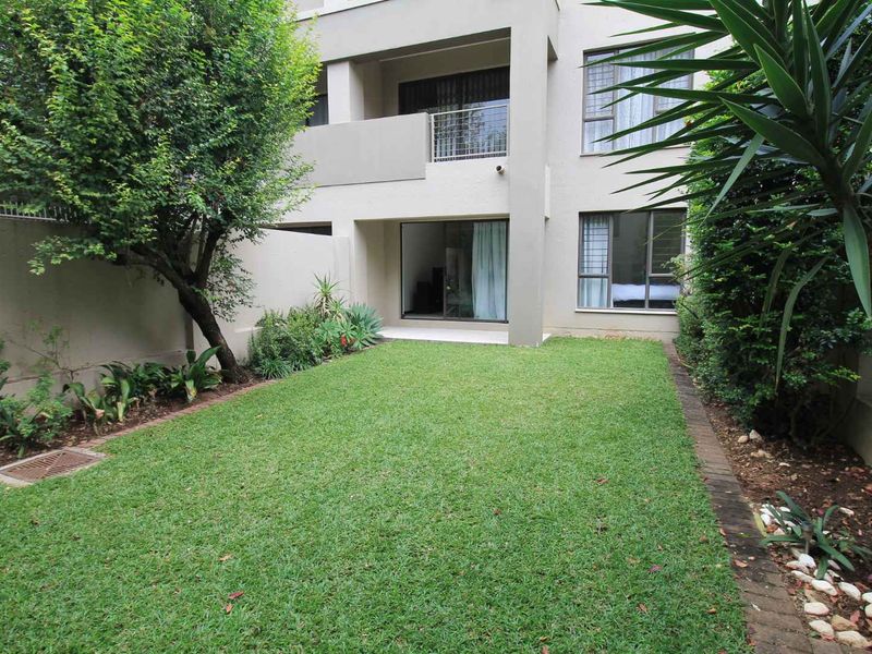 A private rear garden featuring a well-maintained lawn, established landscaping, and direct access to the apartment via sliding glass doors and a balcony.