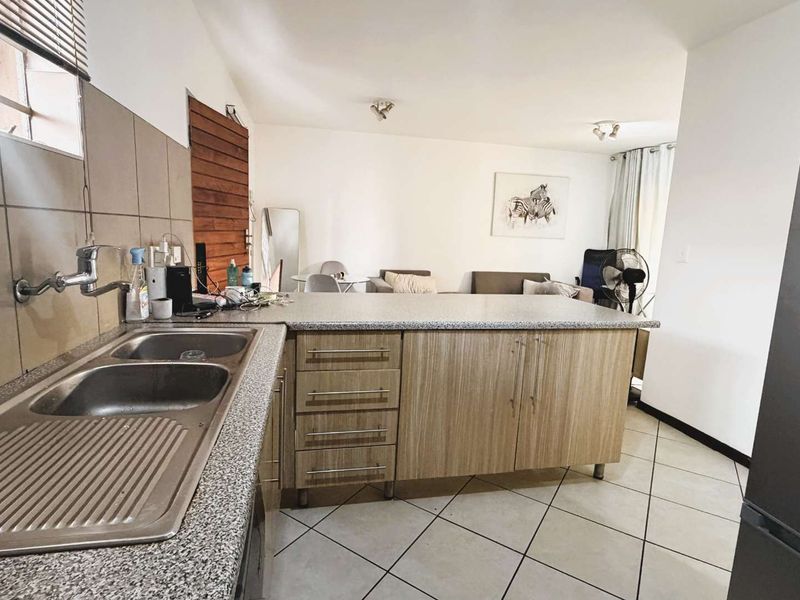 Open-plan kitchen and living area featuring wood-grain cabinetry, granite-look countertops, double basin sink, and neutral ceramic floor tiles.