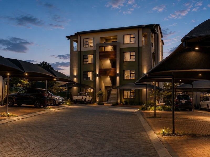 A well-maintained multi-story apartment building at dusk, featuring paved driveway access and covered parking bays with integrated exterior lighting.