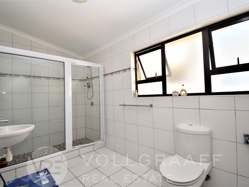 A clean, functional bathroom featuring a glass-enclosed shower, wall-mounted basin, and toilet with neutral white wall and floor tiling.