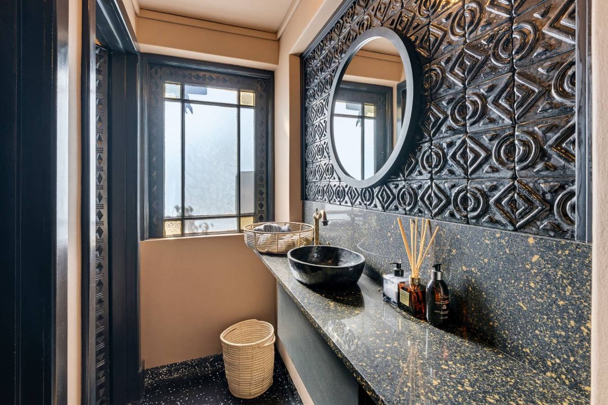 A stylish bathroom featuring a stone vanity countertop, vessel sink, brass faucet, and decorative textured wall tiles with leaded glass window detailing.