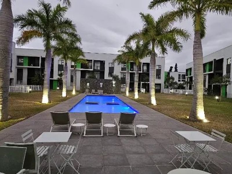 A well-maintained communal swimming pool area surrounded by paved decking, landscaped gardens, and palm trees within an apartment complex.