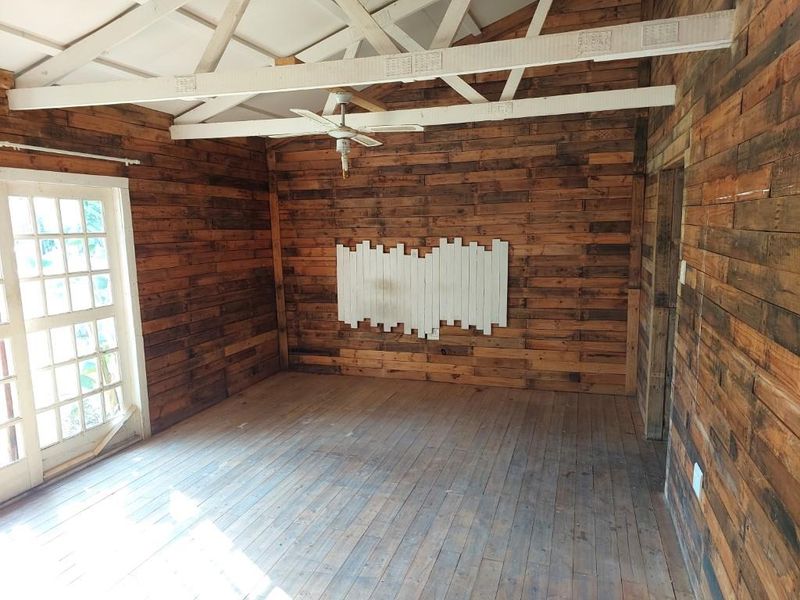 An empty living room featuring rustic timber wall cladding, exposed ceiling beams, a ceiling fan, and classic french doors leading outdoors.