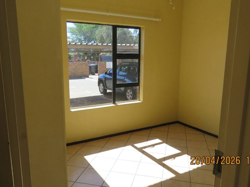 An empty bedroom featuring tiled flooring and a large aluminium-framed window, providing natural light and a view of the exterior parking area.