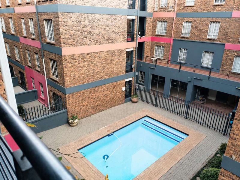 An elevated view of a communal swimming pool area within an apartment complex, featuring brick paving and secure metal perimeter fencing.