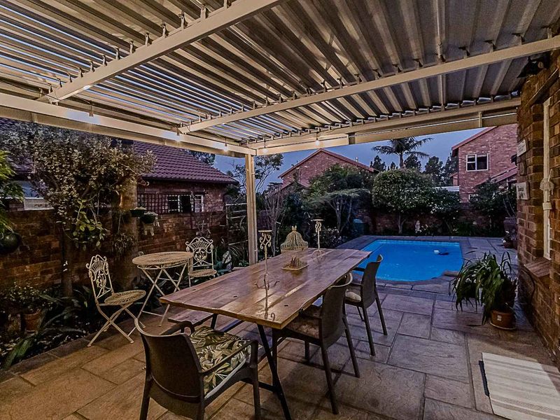 A covered outdoor patio area featuring paved flooring and a corrugated roof, overlooking a private swimming pool and garden space.