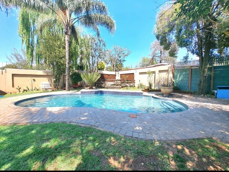 A well-maintained outdoor pool area featuring a paved surround, established garden landscaping, and secure perimeter walling.