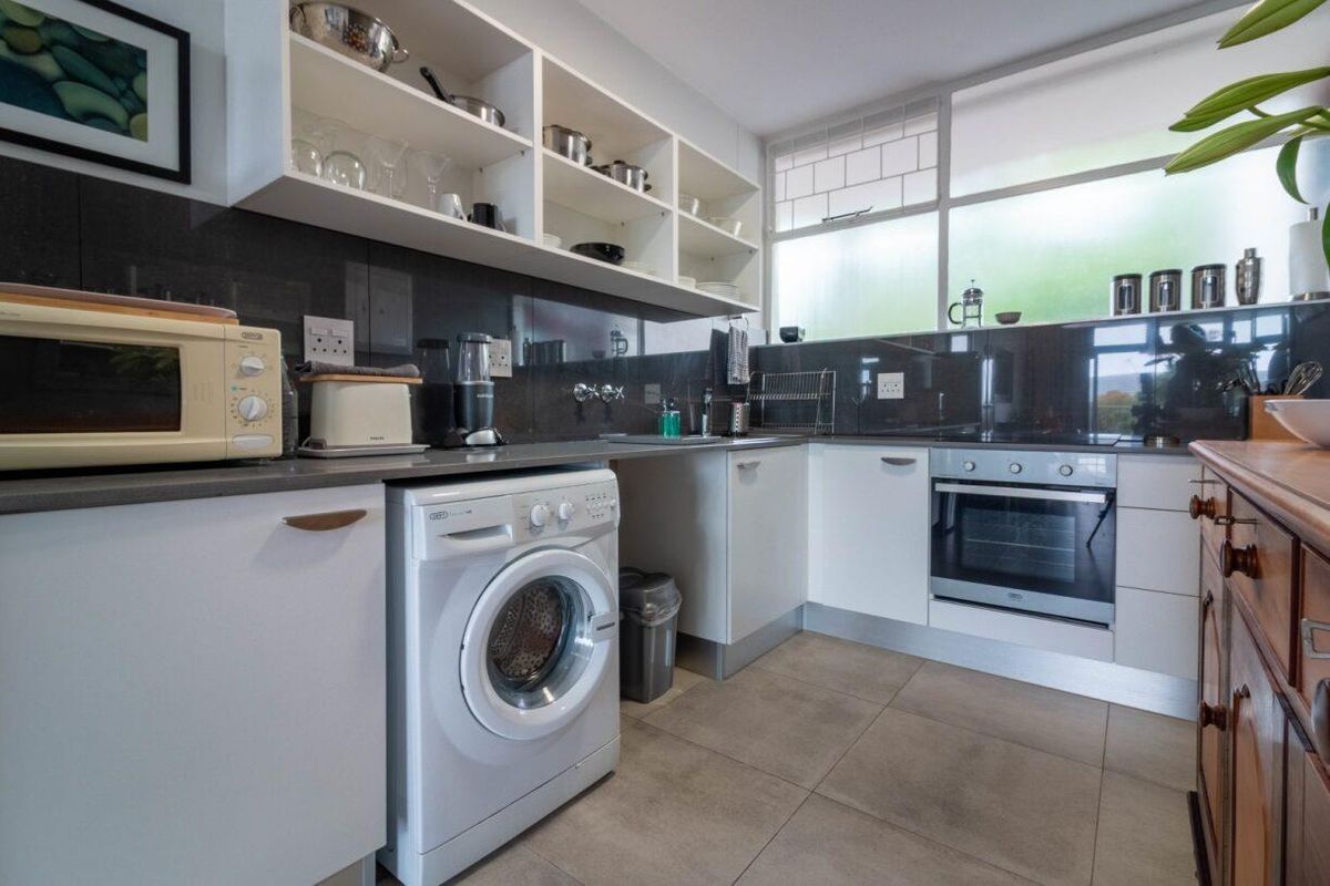 A functional kitchen featuring white cabinetry, open shelving, dark tiled splashbacks, built-in oven, and integrated laundry space with tiled flooring.